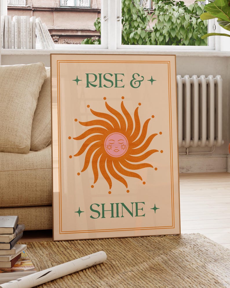 May include: A framed art print with the words "RISE & SHINE" in green, with a stylized orange sun in the center. The sun has a pink face and is surrounded by orange rays. The print is in a wooden frame.
