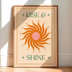 May include: A framed art print with a beige background and a stylized sun illustration in orange and pink. The sun has a smiling face. The words "RISE & SHINE" are printed in green with star accents.