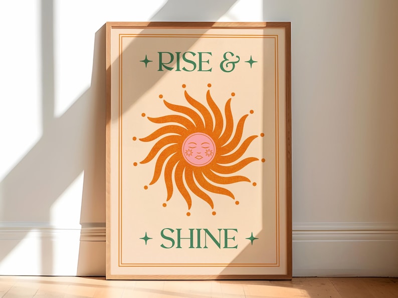 May include: A framed art print with a beige background and a stylized sun illustration in orange and pink. The sun has a smiling face. The words "RISE & SHINE" are printed in green with star accents.