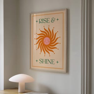 May include: A framed art print featuring a stylized orange sun with a smiling face and radiating lines. The words "RISE &" and "SHINE" are in green text with star accents. The artwork is in a light wood frame.