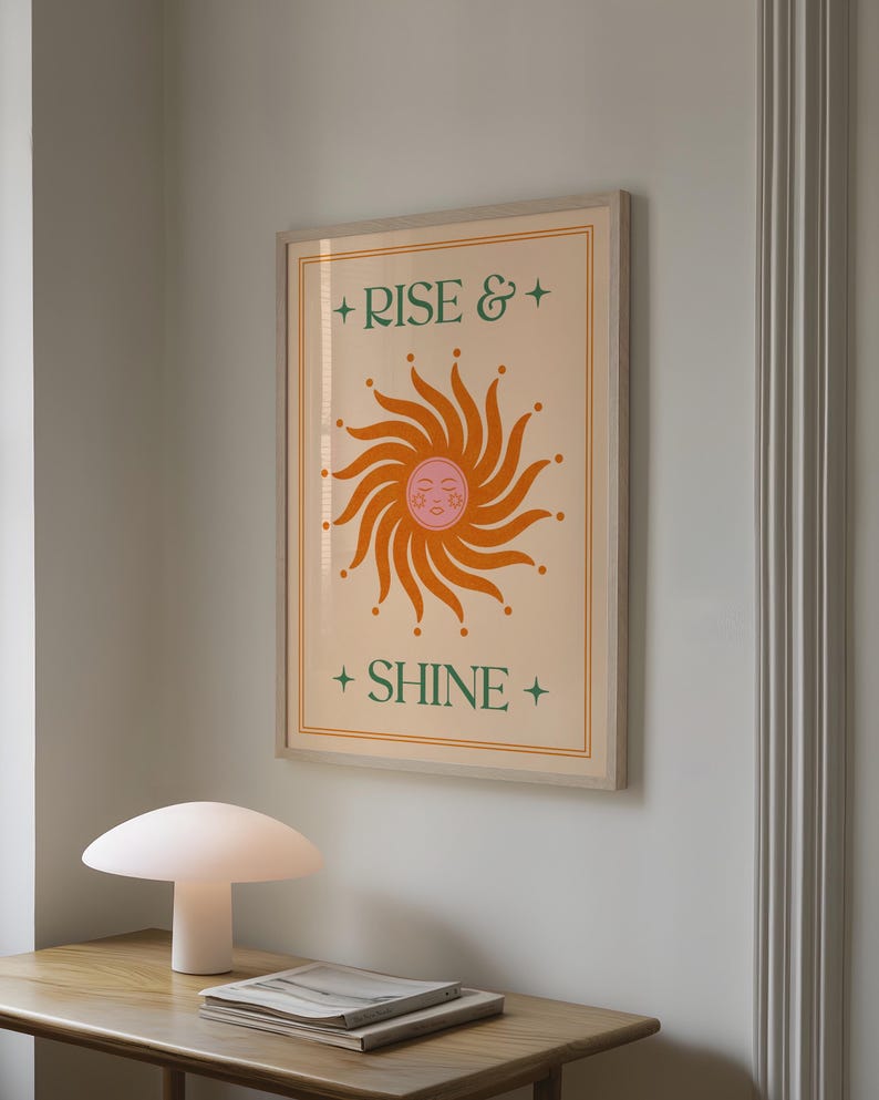 May include: A framed art print featuring a stylized orange sun with a smiling face and radiating lines. The words "RISE &" and "SHINE" are in green text with star accents. The artwork is in a light wood frame.