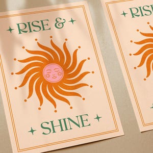 May include: Two art prints with a peach background and a retro sun design. The sun is orange with a pink face. The words "RISE & SHINE" are in green text. The prints have a gold border.