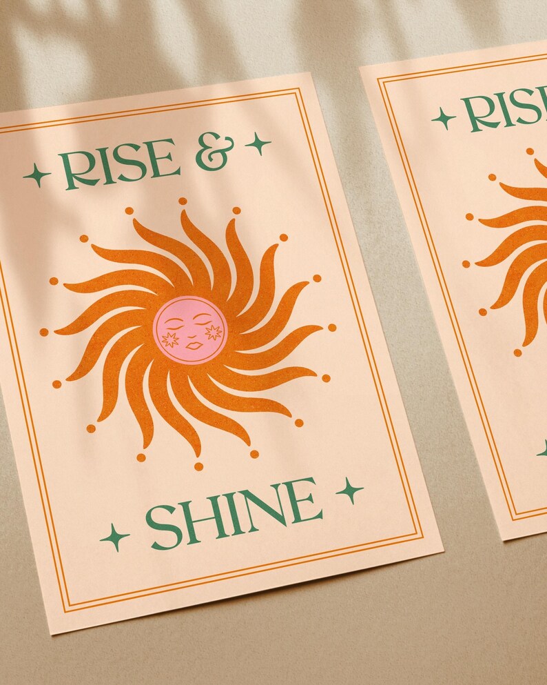 May include: Two art prints with a peach background and a retro sun design. The sun is orange with a pink face. The words "RISE & SHINE" are in green text. The prints have a gold border.