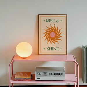 May include: A pink metal side table with a round, glowing lamp and a framed poster that reads "Rise & Shine." The poster features a sun design in orange and green. A silver audio device and books are on the lower shelf.