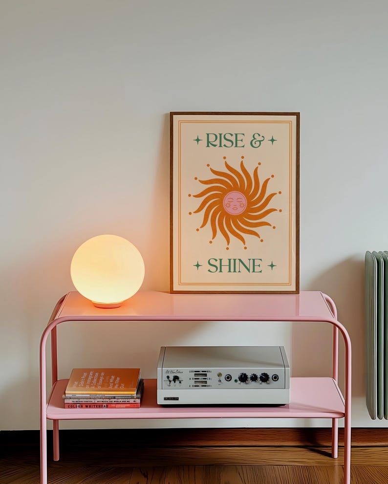 May include: A pink metal side table with a round, glowing lamp and a framed poster that reads "Rise & Shine." The poster features a sun design in orange and green. A silver audio device and books are on the lower shelf.