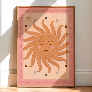 May include: A framed art print depicting a stylized sun with closed eyes and star-shaped cheeks. The sun is a warm orange colour, set against a light pink background with decorative borders and accents in pink and brown. The artwork is displayed in a wooden frame.