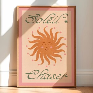 May include: A framed art print featuring a stylized sun illustration with a smiling face and radiating lines. The print has the words "Soleil Chaser" in green script. The artwork is set against a light peach background with a pink border.