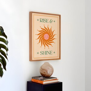 May include: A framed art print with the words "RISE & SHINE" and a sun illustration in orange and pink. The print is in a wooden frame and hangs on a white wall. A decorative vase and books are below the print.