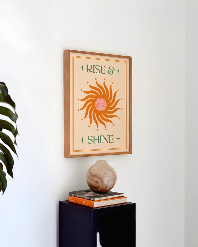 May include: A framed art print with the words "RISE & SHINE" and a sun illustration in orange and pink. The print is in a wooden frame and hangs on a white wall. A decorative vase and books are below the print.