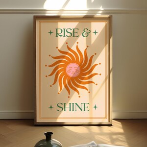 May include: A framed art print with the words "RISE & SHINE" and a stylized sun illustration. The sun is orange with a pink face and surrounded by rays. The print is in a wooden frame and leans against a wall.