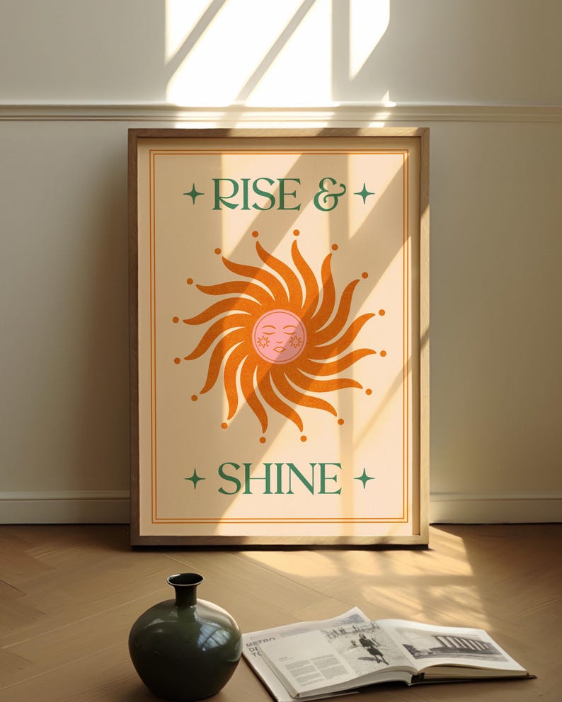 May include: A framed art print with the words "RISE & SHINE" and a stylized sun illustration. The sun is orange with a pink face and surrounded by rays. The print is in a wooden frame and leans against a wall.