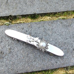 May include: Silver-toned barrette with a decorative floral and pearl accent. The barrette features a central pearl surrounded by an ornate, openwork design. A floral motif adorns the side, adding a touch of nature-inspired elegance to this hair accessory.