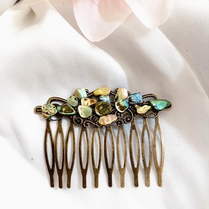 Op de afbeelding: Een bronzen haarspeld met een decoratieve bovenkant met kleurrijke, iriserende stenen. De kam heeft meerdere tanden en is ontworpen om haar vast te zetten. De stenen zijn in tinten groen, blauw en geel.