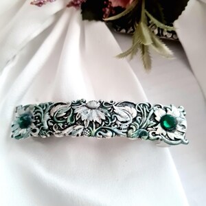 May include: Silver-toned barrette with a floral design and two green gemstone accents. The barrette features detailed relief work of flowers and leaves, with a distressed, vintage appearance. The barrette is approximately 3 inches long.