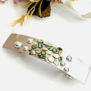 May include: A silver hair clip with a decorative design of iridescent green, blue, and white shell pieces. The clip has a curved shape and is accented with silver beads.