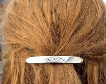 May include: A silver barrette with an ornate, swirling design. The barrette is rectangular and holds back a portion of the person's light brown hair. The hair is wavy and has a natural texture.