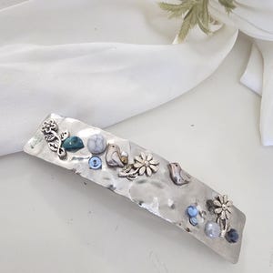 May include: A silver-toned barrette with a hammered texture, adorned with decorative elements. The barrette features small metal flowers, birds, and various blue and gray beads. A stylish hair accessory.