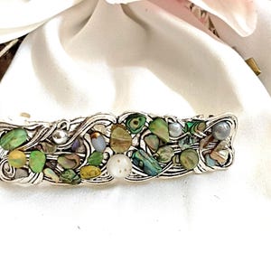 Może przedstawiać: Srebrna spinka do włosów o skomplikowanym, płynnym wzorze. Spinka jest ozdobiona mozaiką kolorowych, nieregularnych kamieni w odcieniach zieleni, brązu i bieli. Kilka małych, okrągłych, srebrnych koralików jest również wkomponowanych w projekt.