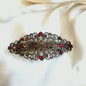 Könnte beinhalten: Antike bronzefarbene Haarspange mit roten Edelsteinakzenten. Die Spange hat ein filigranes Design mit floralen und scrollartigen Details, vor weißem Hintergrund. Ein dekoratives Haaraccessoire.