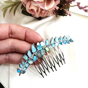 Op de afbeelding: Een zilverkleurige haar kam met een blad ontwerp, versierd met blauwe en turquoise stenen. De kam is geplaatst tegen een achtergrond van roze rozen en witte stof.