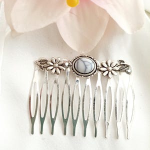 May include: A silver-toned hair comb with a central oval-shaped stone in white and gray. The comb features floral and leaf accents. The comb's tines are evenly spaced and extend downward. The comb is designed for hair styling and is suitable for various occasions.