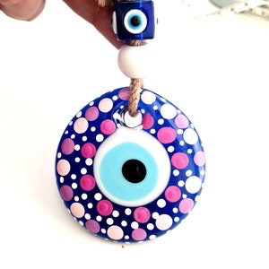 May include: Blue and white evil eye charm with pink and white polka dots. The charm is hanging from a string of white beads and a blue evil eye bead.