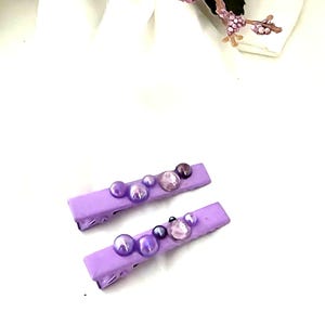 Op de afbeelding: Twee rechthoekige haarclips in een effen lichtpaarse kleur. Elke clip is versierd met een assortiment ronde kralen en gefacetteerde edelstenen in verschillende tinten paars, variërend van licht lavendel tot diep violet, gerangschikt op de bovenkant.