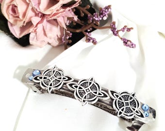 May include: Silver-toned hair barrette with three Celtic knot designs. The barrette features small blue beads and is set against a white background with pink flowers.