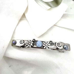 May include: A silver-toned metal hair barrette featuring two daisy-shaped charms, two spiral accents, and three round cabochon stones in shades of blue and grey. The decorative elements are arranged linearly along the length of the hair clip.