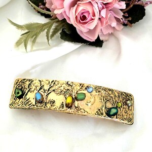 May include: A rectangular gold-toned metal hair clip featuring an embossed forest scene with trees and a crescent moon. The surface is decorated with small, colorful, oval-shaped beads in shades of blue, green, and yellow embedded in the metal.