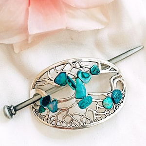May include: Silver hair stick with an oval, openwork design, decorated with turquoise-coloured stones. The stick has a decorative end and a long, slender pin for securing hair. A pink flower is in the background.