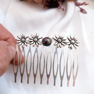 May include: Silver hair comb featuring daisy flower accents and a central, iridescent bead. The comb has multiple tines for securing in hair. A decorative hair accessory.