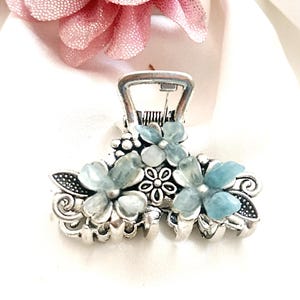 May include: Silver-toned metal hair claw clip adorned with light blue flower-shaped accents. The clip features a decorative floral design with intricate details and a secure clasp, perfect for various hairstyles.