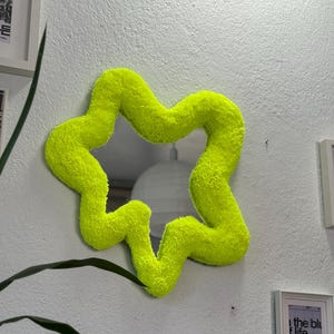 May include: A neon green, star-shaped mirror with a fluffy, textured frame. The mirror is mounted on a white wall, with framed pictures nearby. The mirror's unique shape and bright colour make it a decorative accent.