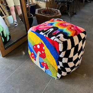 May include: A colourful, square-shaped floor cushion with a rainbow, checkered, and mushroom design. The cushion is made of a soft, plush fabric.