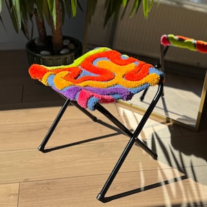 May include: A folding stool with a black metal frame and a tufted seat featuring a vibrant, multi-colored abstract pattern in shades of orange, red, yellow, blue, and purple. The textured seat surface has a raised, plush, yarn-like appearance.