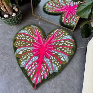May include: A heart-shaped rug with a pink and green leaf design. The rug is made of a soft, plush material and has a white background.