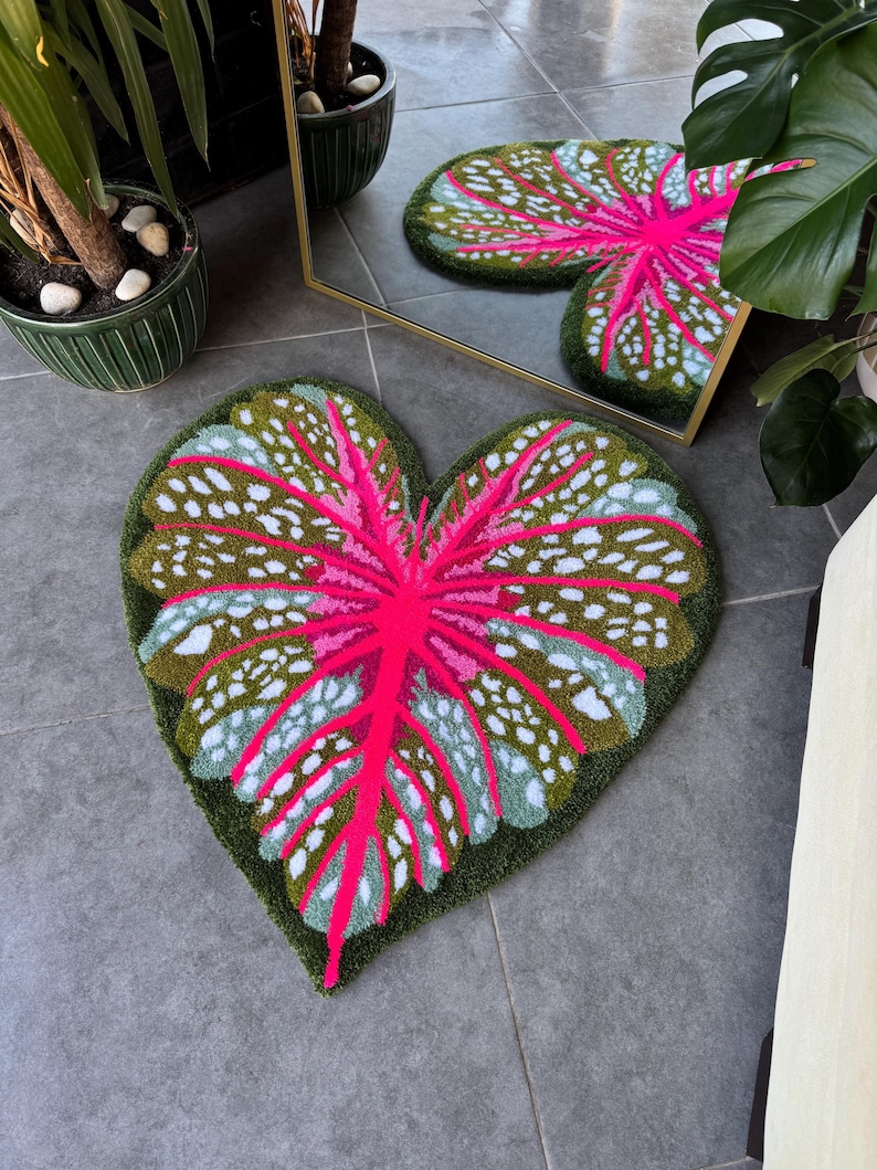 May include: A heart-shaped rug with a pink and green leaf design. The rug is made of a soft, plush material and has a white background.