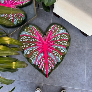 May include: A heart-shaped rug with a pink and green leaf design. The rug is made of a soft, plush material and has a white background with green and pink accents.