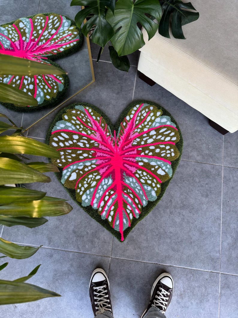 May include: A heart-shaped rug with a pink and green leaf design. The rug is made of a soft, plush material and has a white background with green and pink accents.