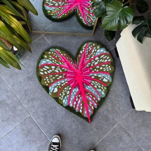 May include: A pink and green heart-shaped rug with a leaf design. The rug is made of a soft, plush material and has a white border.