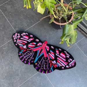 Puede incluir: Una alfombra negra con forma de mariposa con rayas rosas, moradas, azules y amarillas. La alfombra está sobre un suelo de baldosas grises.