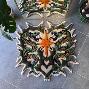 Könnte beinhalten: Ein getufteter Teppich mit einem symmetrischen Design aus zwei grünen Schlangen mit orangefarbenen und blauen Schuppen, die einem zentralen orangefarbenen Stern gegenüberstehen. Der Teppich hat einen cremefarbenen Hintergrund, dunkelgrüne gezackte Ränder und ein herzförmiges Motiv am unteren Rand.