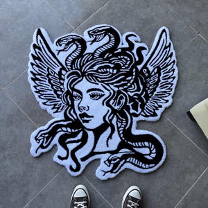 Könnte beinhalten: Ein getufteter Teppich mit einer schwarz-weißen grafischen Illustration einer Medusa-Figur mit Schlangen als Haaren und großen Flügeln an den Seiten. Das Design ist in einem kontrastreichen, kräftigen Linienkunststil mit texturierter Oberfläche.