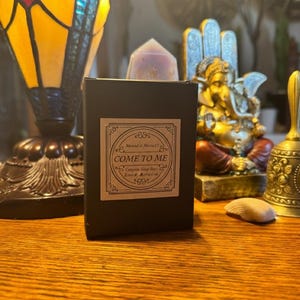 May include: A black box with a label that reads "COME TO ME" and "Love & Attraction". A crystal point sits atop the box. The box is on a wooden surface, with a stained glass lamp and a gold statue in the background.