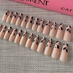 May include: A set of 20 long, almond-shaped, fake nails with a cow print design. The nails are a light pink color with black spots.