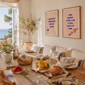Puede incluir: Una mesa de comedor preparada para una comida con vistas al mar. La mesa está puesta con platos, vasos y un mantel a rayas. Dos cuadros enmarcados con citas sobre el vino cuelgan de la pared. El texto de los cuadros dice: "I raise my vibration and my glass" y "Good energy flows like a full glass of wine."