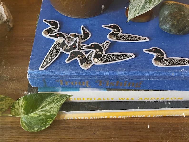 Puede incluir: Una pila de libros con una cubierta azul que presenta varios parches bordados de p&aacute;jaros loon. El libro superior tiene el t&iacute;tulo "Pesca de truchas" impreso en el lomo.