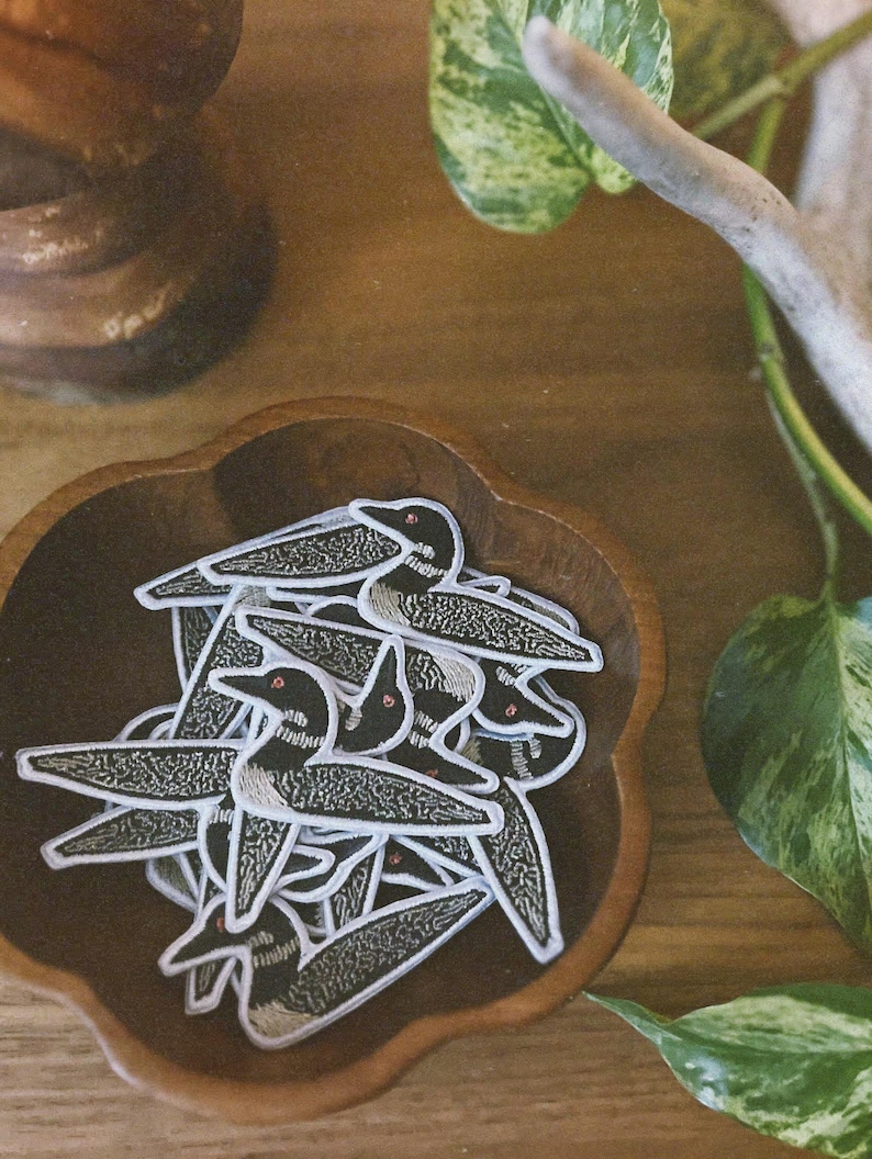 May include: A wooden bowl filled with black and white embroidered loon bird patches. The patches have red eyes and are arranged in a pile.