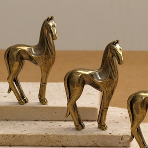 Könnte beinhalten: Drei bronzefarbene Pferdfiguren sind auf einer mehrstufigen, hellfarbigen Oberfläche angeordnet. Die Pferde stehen, mit detaillierten Mähnen und schlanken Beinen. Der Hintergrund ist in einem neutralen Beigeton gehalten.